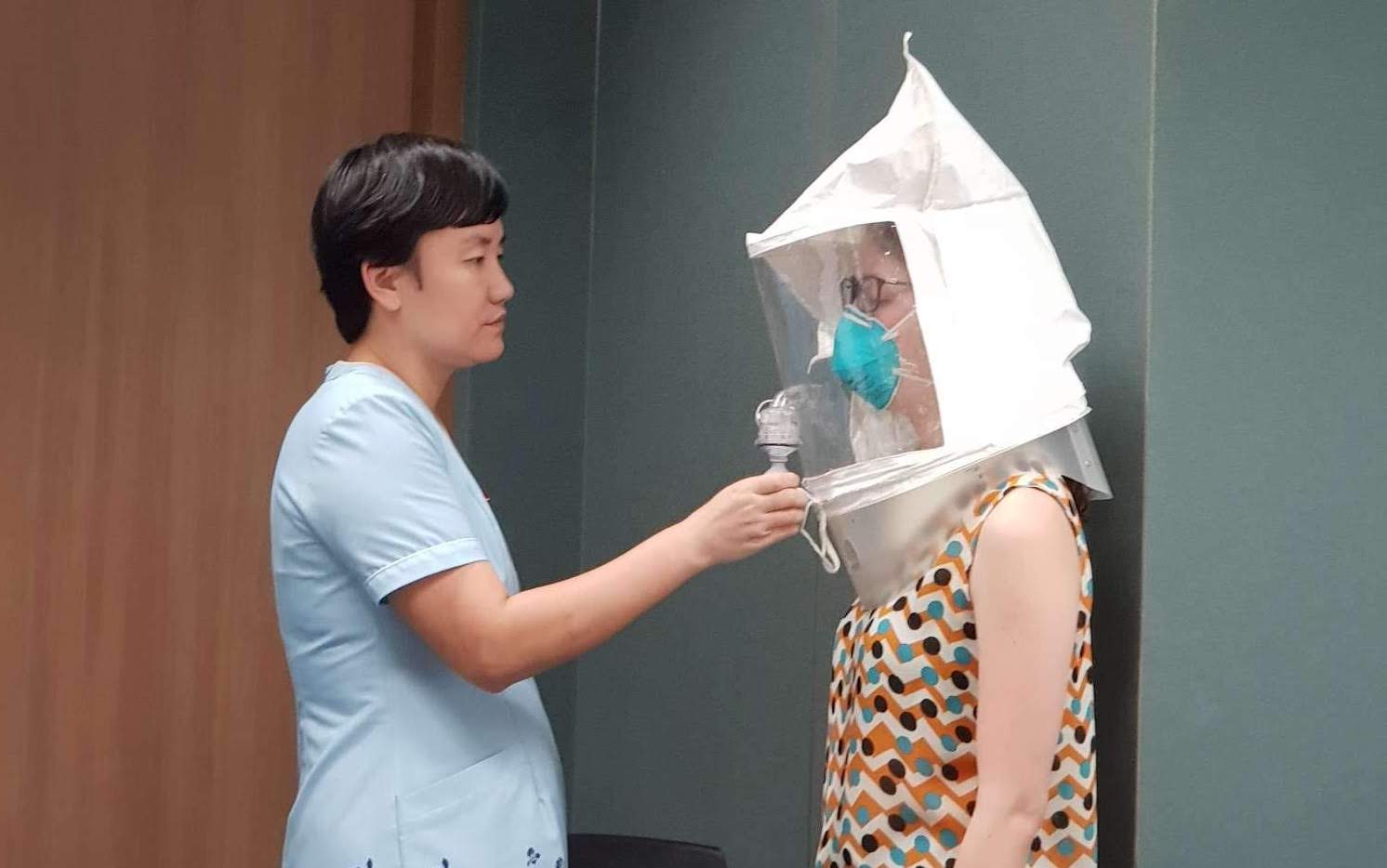 Two women stand facing each other. One women administers a spray to the other which wears an enclosed bonnet and N95 mask, to ensure proper fitting of N95 masks