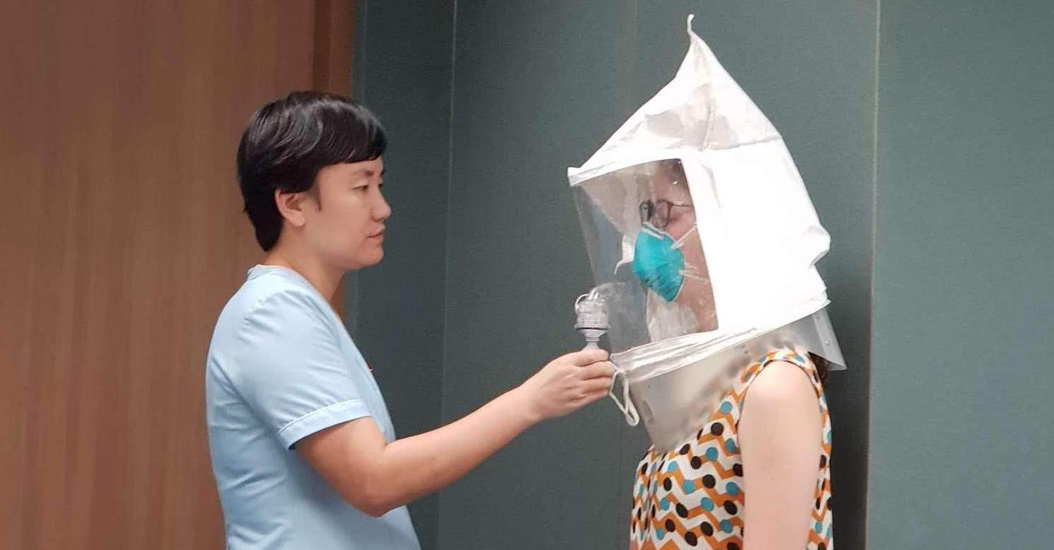Two women stand facing each other. One women administers a spray to the other which wears an enclosed bonnet and N95 mask, to ensure proper fitting of N95 masks